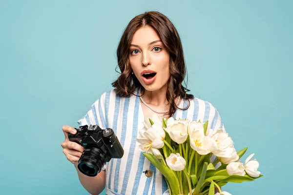 Excited young woman holding digital camera and bouquet of white tulips isolated on blue — Stock Photo