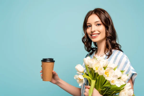 Joyeuse jeune femme tenant du café à emporter et bouquet de tulipes blanches et regardant la caméra isolée sur bleu — Photo de stock