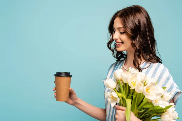 Весела молода жінка тримає каву, щоб піти і букет білих тюльпанів ізольовані на синьому — стокове фото