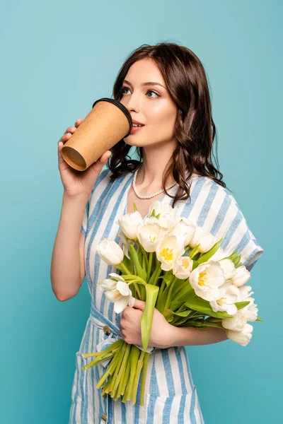 Soñadora joven bebiendo café para llevar mientras sostiene tulipanes blancos aislados en azul - foto de stock