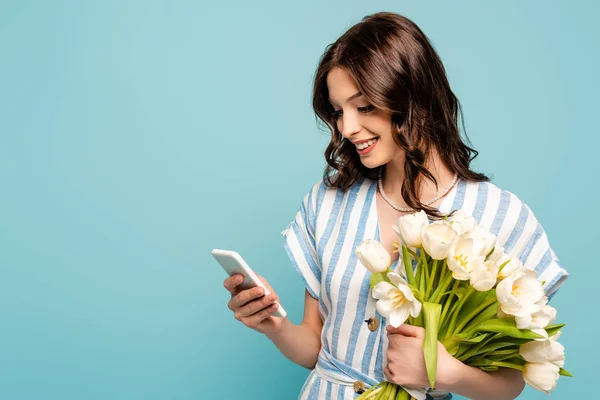 Felice giovane donna che chatta su smartphone mentre tiene tulipani bianchi isolati su blu — Foto stock