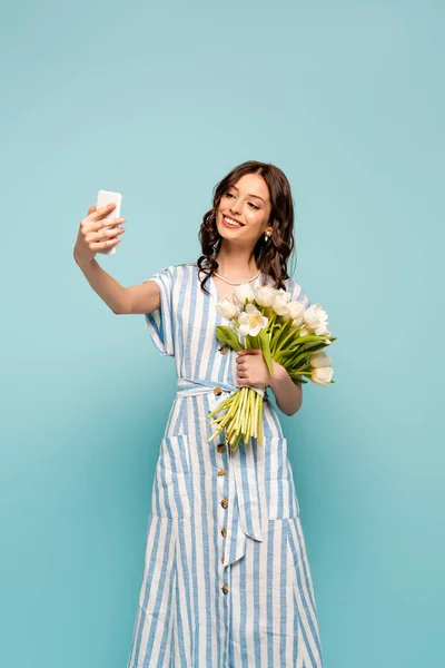 Приваблива молода жінка бере селфі на смартфон, тримаючи білі тюльпани ізольовані на синьому — стокове фото