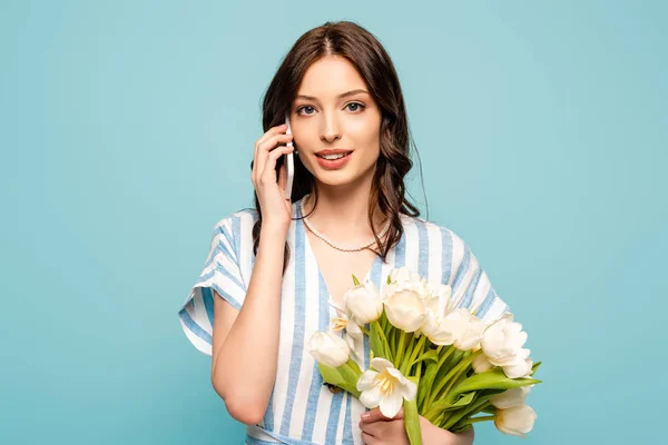 Belle jeune femme parlant sur smartphone tout en tenant des tulipes blanches isolées sur bleu — Photo de stock