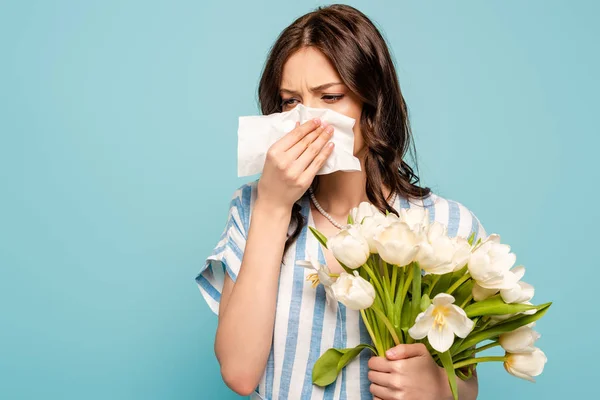 Mujer enferma limpiando la nariz con servilleta de papel mientras sostiene tulipanes blancos aislados en azul - foto de stock