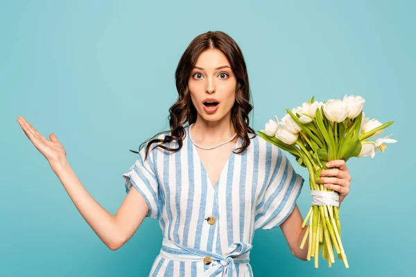 Scioccata giovane donna che tiene bouquet di tulipani mentre in piedi con il braccio aperto isolato su blu — Foto stock