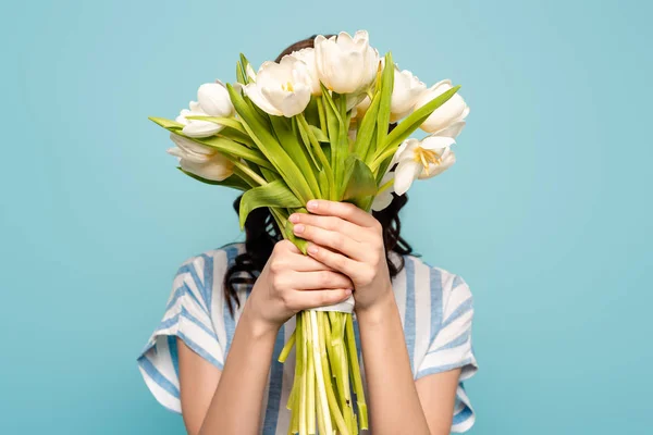 Joven mujer oscureciendo la cara con ramo de tulipanes blancos aislados en azul - foto de stock