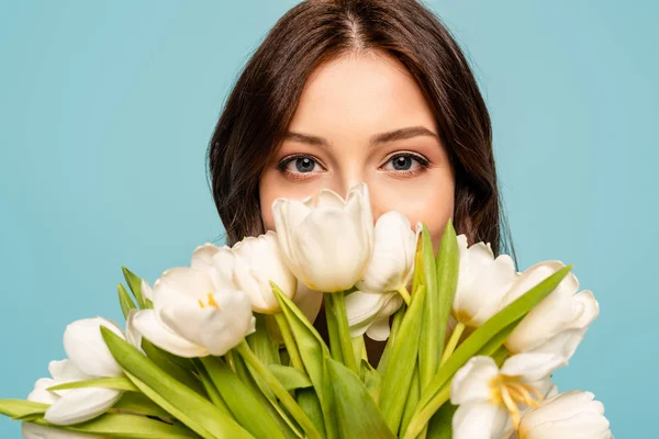 Hermosa mujer joven disfrutando del sabor de los tulipanes blancos mientras mira la cámara aislada en azul - foto de stock