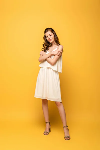 Full length view of beautiful young woman smiling at camera while hugging herself on yellow background — Stock Photo