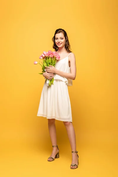 Ganzkörperansicht der schönen jungen Frau lächelt in die Kamera, während sie einen Strauß rosa Tulpen auf gelbem Hintergrund hält — Stockfoto