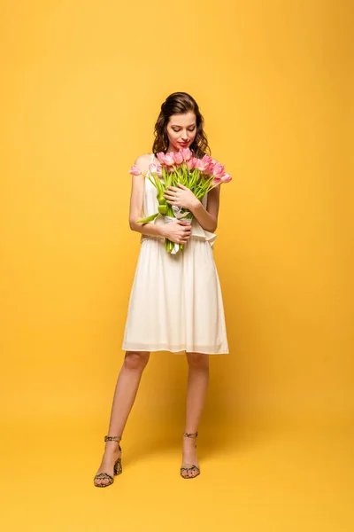 Full length view of attractive young woman smiling while holding bouquet of pink tulips on yellow background — Stock Photo