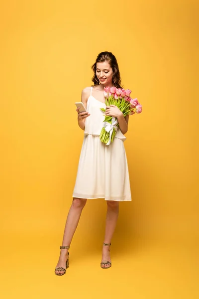 Piena vista lunghezza di felice giovane donna che chiacchiera su smartphone tenendo in mano mazzo di tulipani rosa su sfondo giallo — Foto stock