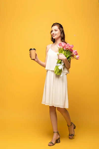 Full length view of young woman smiling while holding bouquet of pink tulips and coffee to go on yellow background — Stock Photo