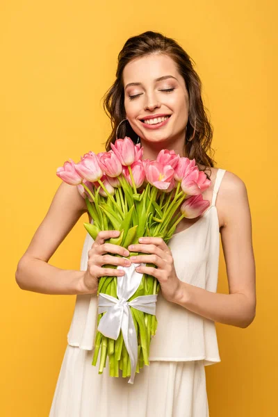 Happy young woman holding bouquet of pink tulips while smiling with closed eyes isolated on yellow — Stock Photo