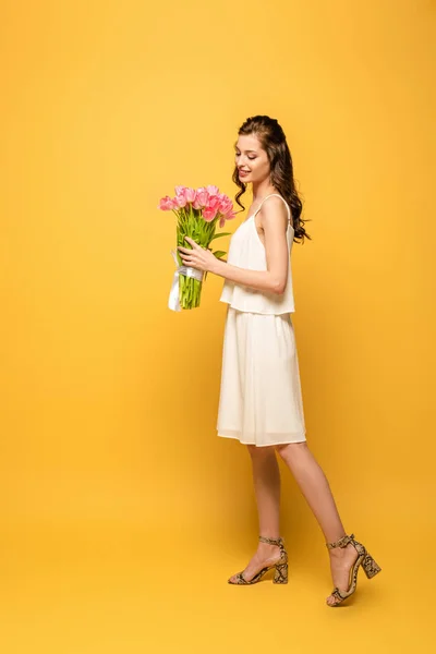 Full length view of smiling young woman holding bouquet of pink tulips on yellow background — Stock Photo