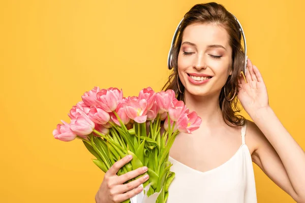 Happy young woman holding bouquet of pink tulips while listening music in wireless headphones with closed eyes isolated on yellow — Stock Photo