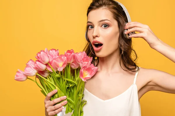 Schockierte junge Frau mit drahtlosen Kopfhörern, die in die Kamera schaut, während sie einen Strauß rosa Tulpen auf gelb isoliert hält — Stockfoto