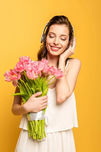 Happy young woman holding bouquet of pink tulips while listening music in wireless headphones with closed eyes isolated on yellow — Stock Photo