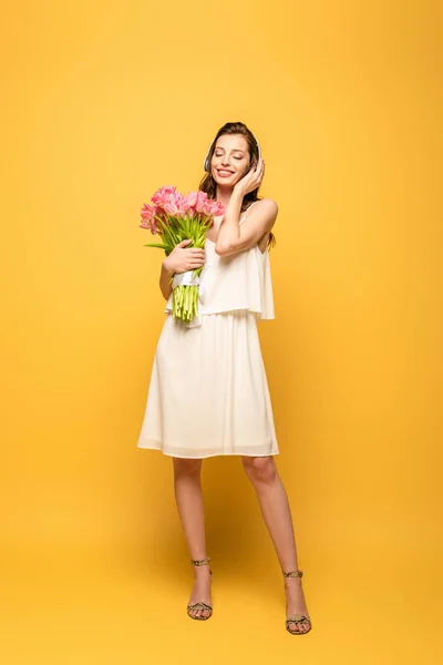 Full length view of happy young woman in wireless headphones holding bouquet of pink tulips on yellow background — Stock Photo