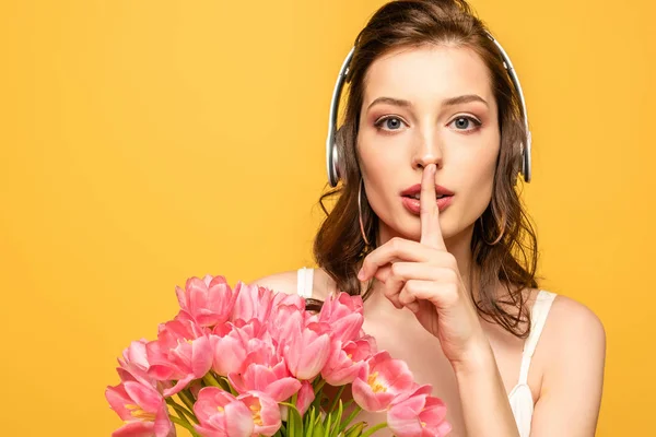 Attractive young woman in wireless headphones showing hush gesture while holding bouquet of tulips isolated on yellow — Stock Photo