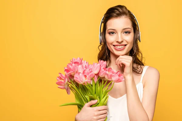 Jolie jeune femme en casque sans fil souriant à la caméra tout en tenant bouquet de tulipes isolées sur jaune — Photo de stock