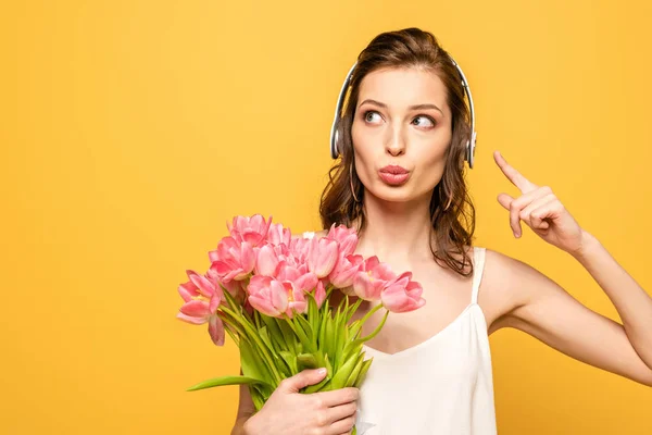 Mujer joven reflexiva en auriculares inalámbricos apuntando con el dedo mientras sostiene ramo de tulipanes aislados en amarillo - foto de stock