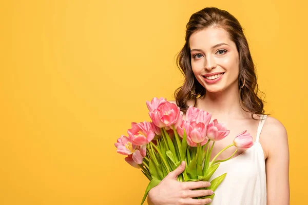 Alegre joven mujer sonriendo a la cámara mientras sostiene ramo de tulipanes rosados aislados en amarillo - foto de stock