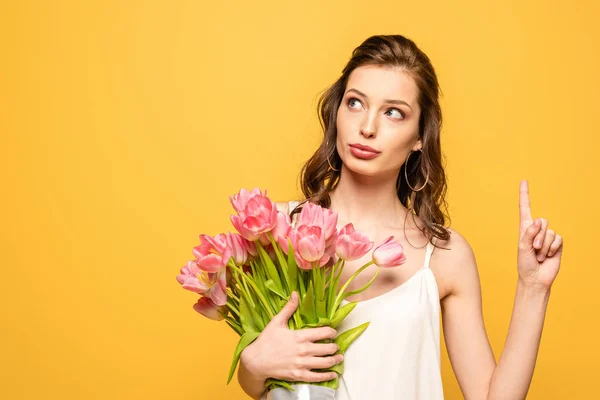 Mujer joven reflexiva mirando hacia otro lado mientras sostiene ramo de tulipanes rosados aislados en amarillo - foto de stock
