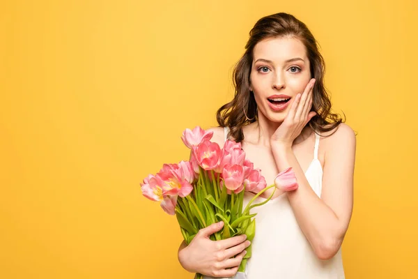 Schockierte junge Frau mit einem Strauß rosa Tulpen und einem anrührenden Gesicht, während sie isoliert auf gelb in die Kamera blickt — Stockfoto