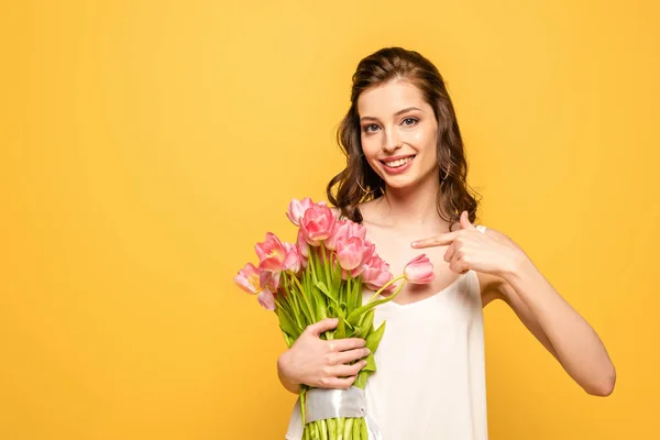 Smiling young woman pointing with finger at bouquet of pink tulips while looking at camera isolated on yellow — Stock Photo