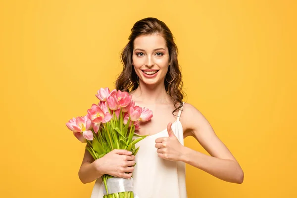 Happy young woman holding bouquet of pink tulips while looking at camera and showing thumb up isolated on yellow — Stock Photo