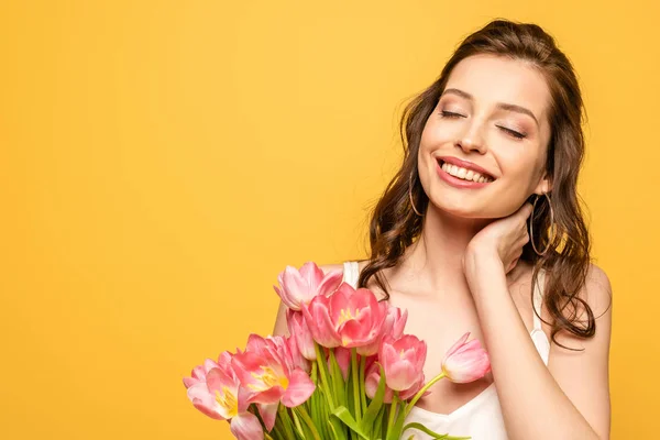Felice giovane donna che tiene bouquet di tulipani rosa mentre tocca il collo con gli occhi chiusi isolati sul giallo — Foto stock