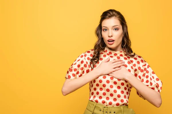 Überraschte junge Frau hält die Hände auf der Brust, während sie in die Kamera schaut, isoliert auf gelb — Stockfoto