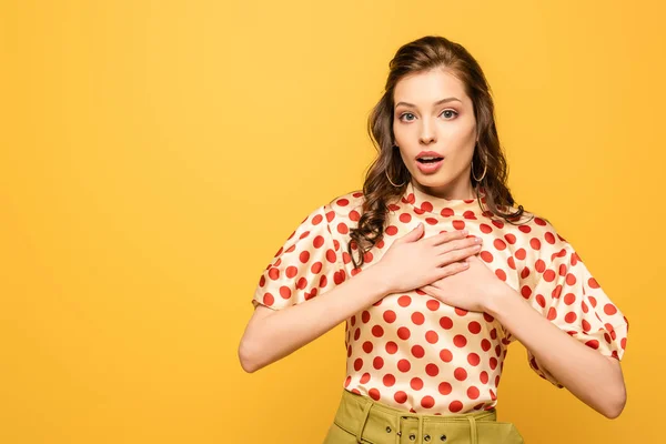 Sorprendida mujer joven cogida de la mano en el pecho mientras mira a la cámara aislada en amarillo - foto de stock