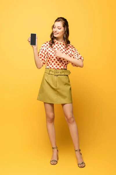 Ganzkörperansicht attraktiver junger Frau, die lächelt, während sie mit dem Finger auf Smartphone mit leerem Bildschirm auf gelbem Hintergrund zeigt — Stockfoto