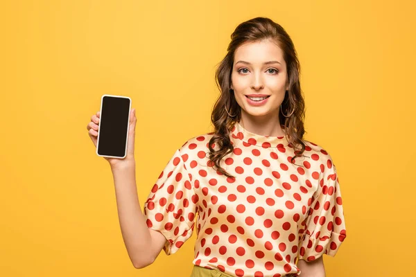 Красива молода жінка посміхається на камеру, показуючи смартфон з порожнім екраном ізольовано на жовтому — стокове фото