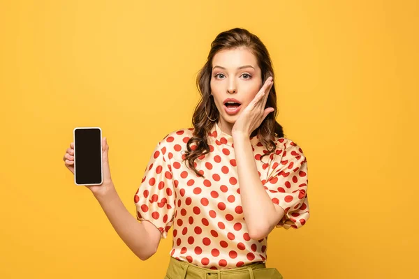 Шокована молода жінка торкається обличчя, показуючи смартфон з порожнім екраном ізольовано на жовтому — стокове фото