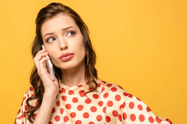 Mujer joven reflexiva mirando hacia otro lado mientras habla en el teléfono inteligente aislado en amarillo - foto de stock