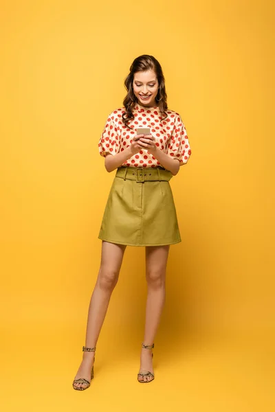 Full length view of happy young woman chatting on smartphone on yellow background — Stock Photo