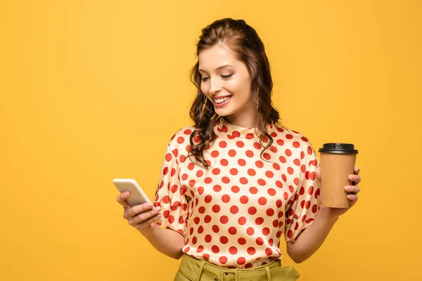 Щаслива молода жінка тримає каву, щоб піти, використовуючи смартфон ізольовано на жовтому — стокове фото