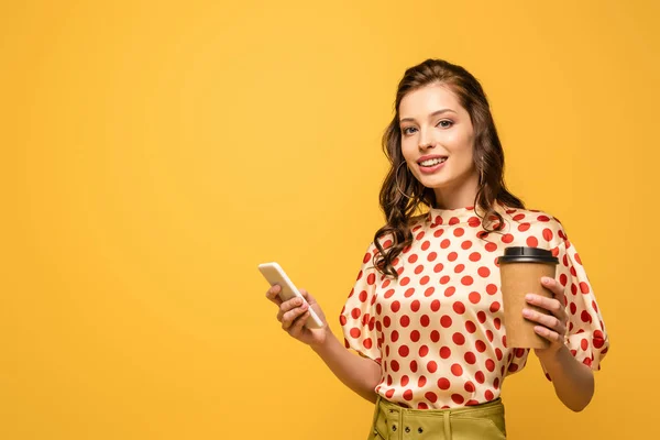 Attractive young woman holding coffee to go while using smartphone and looking at camera isolated on yellow — Stock Photo