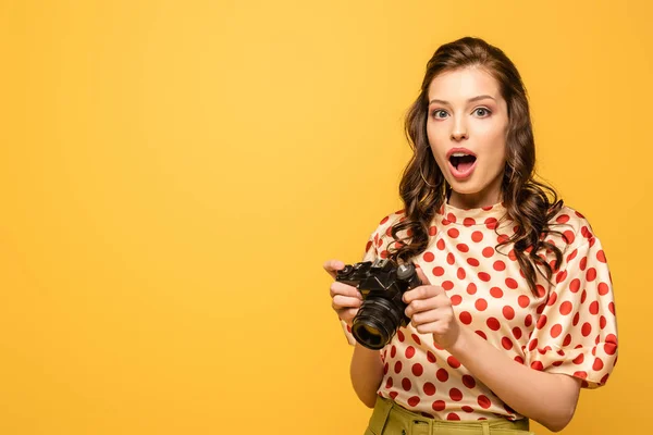 Shocked young woman holding digital camera isolated on yellow — Stock Photo