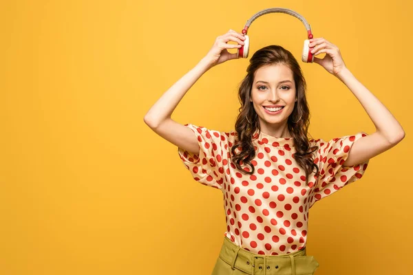 Щаслива молода жінка посміхається на камеру, тримаючи бездротові навушники над головою ізольовані на жовтому — стокове фото