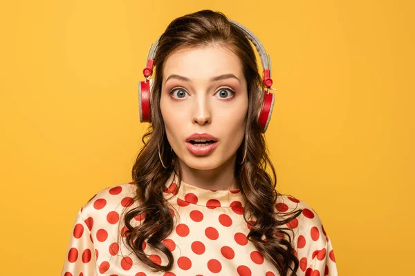 Шокована молода жінка в бездротових навушниках дивиться на камеру ізольовано на жовтому — стокове фото