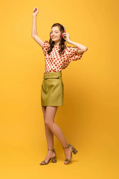 Full length view of happy young woman in wireless headphones dancing with closed eyes on yellow background — Stock Photo