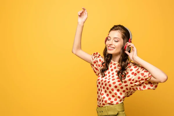 Cheerful young woman in wireless headphones dancing with closed eyes isolated on yellow — Stock Photo