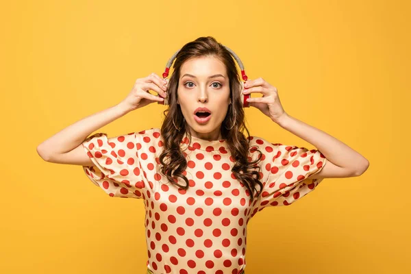 Conmocionó a la joven tocando auriculares inalámbricos mientras miraba a la cámara aislada en amarillo - foto de stock