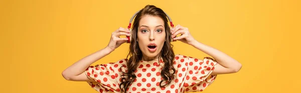 Panoramic shot of shocked young woman touching wireless headphones while looking at camera isolated on yellow — Stock Photo