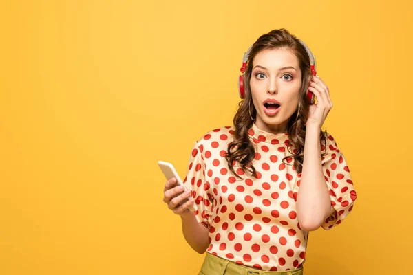 Шокована молода жінка в бездротових навушниках дивиться на камеру, тримаючи смартфон ізольовано на жовтому — стокове фото