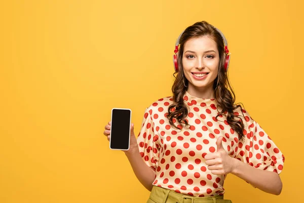 Joyeuse jeune femme dans les écouteurs sans fil tenant smartphone avec écran blanc et montrant pouce vers le haut isolé sur jaune — Photo de stock