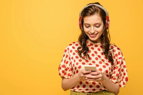 Alegre joven mujer en auriculares inalámbricos charlando en el teléfono inteligente aislado en amarillo — Stock Photo